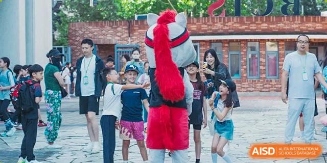 Young students interact excitedly with a person wearing a large grey and red horse mascot costume in an outdoor courtyard during a festive campus celebration at Beijing City International School.