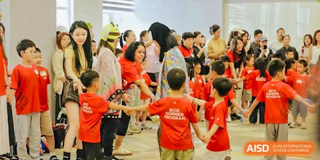 Young students in bright red summer program shirts stand in a circle and hold hands during an organized group activity in a well-lit indoor common area at Beijing City International School.