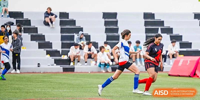 A female student athlete in a white, blue, and red jersey sprints across a green soccer field during a competitive outdoor sports match held at Beijing City International School.
