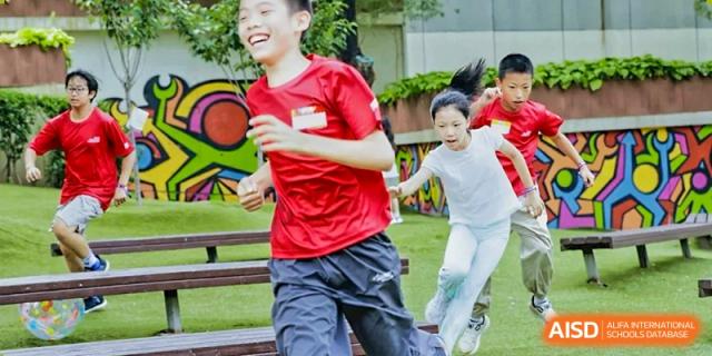 A group of smiling students in red and white shirts runs enthusiastically through an outdoor park area during a high-energy summer program session at Beijing City International School.