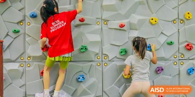 Students in red and beige clothing challenge themselves by climbing up a grey textured indoor rock wall with colorful handholds during a physical activity at Beijing City International School.