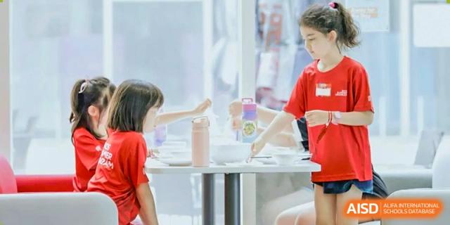 A young student in a red shirt sits at a white table and uses chopsticks to enjoy a meal with classmates during a supervised lunch break at Beijing City International School.