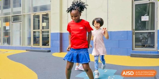 Two young girls in casual summer attire participate in an outdoor game on a playground featuring colorful ground markings and blue walls at Beijing City International School.