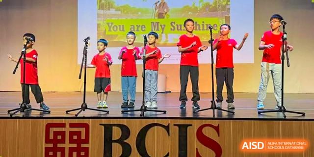 Five students dressed in bright red shirts stand on a stage behind microphones and perform a song entitled You Are My Sunshine for an audience at Beijing City International School.