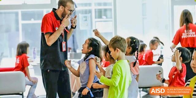 An instructor in a red and black shirt interacts closely with a group of young, curious students during a lively and engaging indoor classroom activity at Beijing City International School.