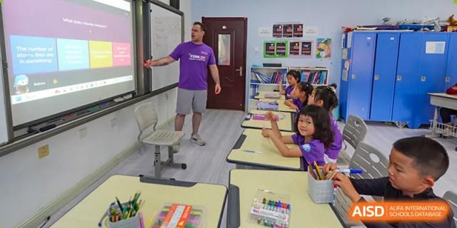 A male teacher in a purple shirt points to a whiteboard while instructing a group of young students at the Canadian International School of Shenyang.