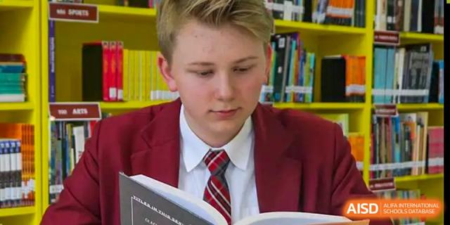 A student wearing a formal red school blazer and tie focuses on reading a book in the library at the Canadian International School of Shenyang.
