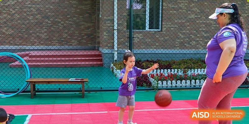 A young girl in a purple shirt practices dribbling a basketball on a red and green outdoor sports court during recess at the Canadian International School of Shenyang.