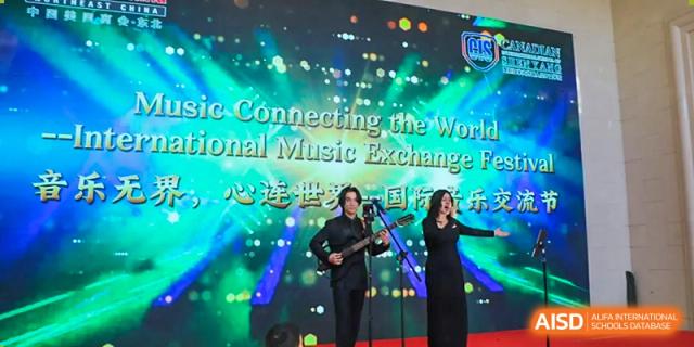 A man plays guitar and a woman sings on a stage with a bright green "International Music Exchange" backdrop at the Canadian International School of Shenyang.