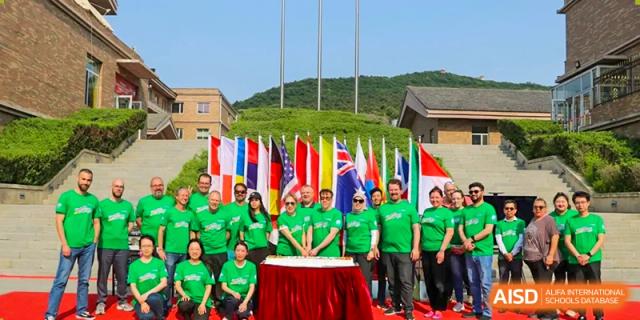 A group of staff members in matching green shirts stands before an international flag display for a cake cutting at the Canadian International School of Shenyang.