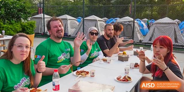 Staff members in green shirts wave while sitting at an outdoor table with food and tents in the background at the Canadian International School of Shenyang.