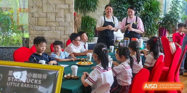 Students sit at a green table with snacks while a staff member in a black apron speaks to them at the Canadian International School of Shenyang.