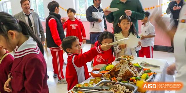 Students in red uniforms excitedly reach for food at a catered buffet event held indoors at the Canadian International School of Shenyang.