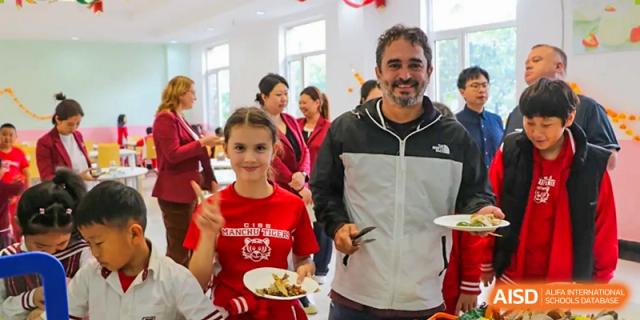 A student in a red shirt and an adult in a grey jacket smile while holding plates of food in the dining area at the Canadian International School of Shenyang.