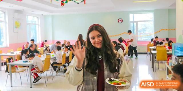 A smiling female student in a plaid jacket waves to the camera while holding a plate of food in the dining hall at the Canadian International School of Shenyang.