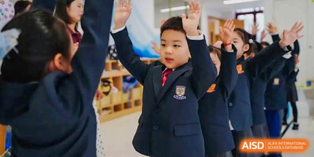 A young boy in a dark uniform suit raising his hand enthusiastically in class, actively participating in a lesson at Concordia International School Ningbo.