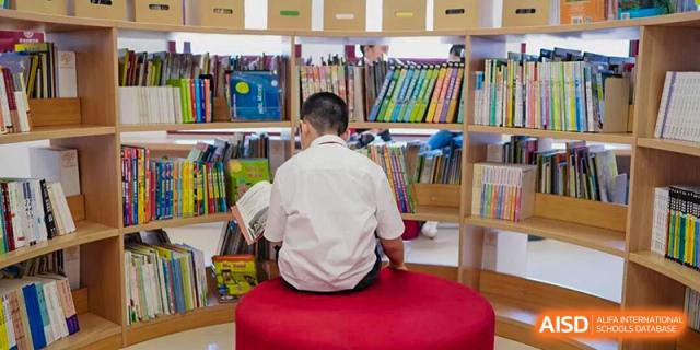 A student sitting on a comfortable red stool, reading a book in front of a colorful bookshelf in the school library at Concordia International School Ningbo.