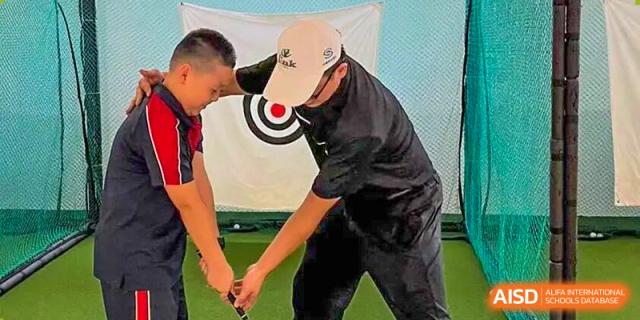 An instructor helping a man practice his swing in an indoor golf training area with a target net visible in the background at Concordia International School Ningbo.