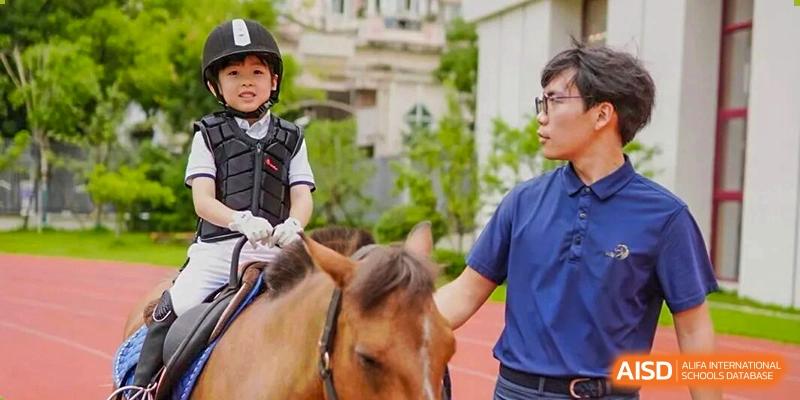 A young student in riding gear sitting on a horse with an instructor or coach nearby during an outdoor equestrian lesson at Concordia International School Ningbo.