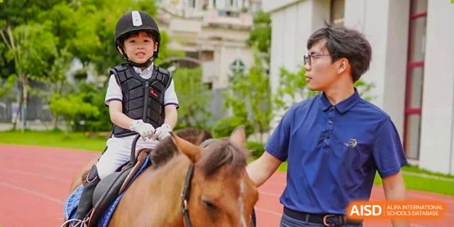 A young student in riding gear sitting on a horse with an instructor or coach nearby during an outdoor equestrian lesson at Concordia International School Ningbo.