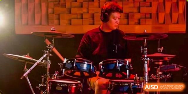 A focused musician playing a drum set with headphones on, illuminated by warm stage lights, possibly during a school performance at Concordia International School Ningbo.