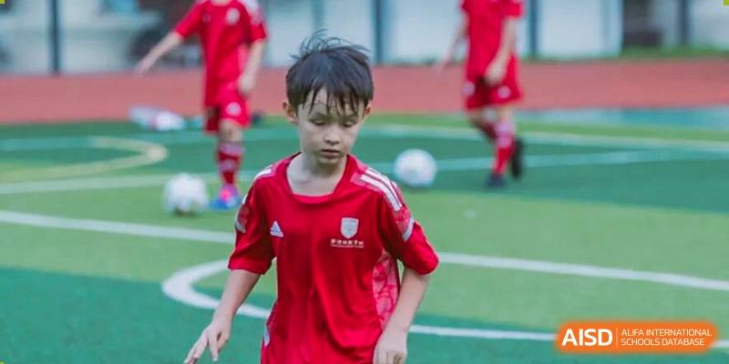A young boy in a red sports uniform playing football/soccer on a field, concentrating on the game during school sports practice at Concordia International School Ningbo.
