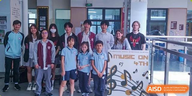 A group of students of various ages stand together next to a "Music Day" poster featuring musical notes and symbols at École Française Internationale de Shenzhen (EFIS).
