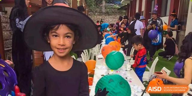 A girl in a witch hat smiles next to a long table of colorful paper lanterns at École Française Internationale de Shenzhen (EFIS).