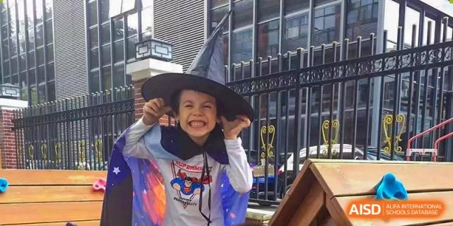 A young student smiling brightly while wearing a black witch hat and a blue cape during a school event at École Française Internationale de Shenzhen(EFIS).