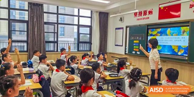 A bright classroom scene showing many students seated at desks, actively engaged and raising their hands to answer questions during a lesson at Hefei Xinhua Academy.