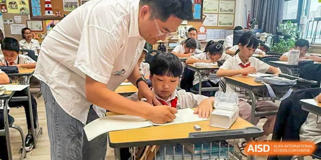 A male teacher leaning over to help a young student with his drawing or writing at a desk during class, Hefei Xinhua Academy.