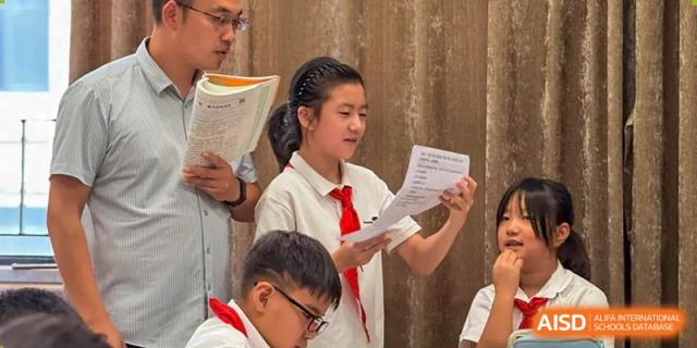 A young female student wearing a white shirt and red tie stands and reads from a paper during a classroom activity while a teacher observes nearby at Hefei Xinhua Academy.