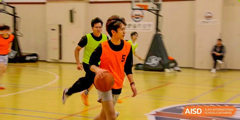 Two young male students playing basketball indoors, one wearing a bright orange pinnie and dribbling, the other in a neon green pinnie, during a game or practice at Hefei Xinhua Academy.
