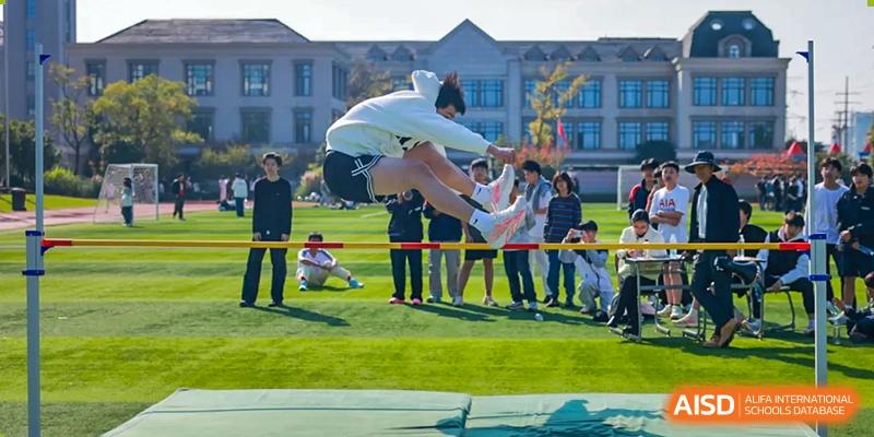 A student wearing a white hoodie performing a high jump over a bar on an athletic field, landing onto a green crash mat, cheered on by others at Hefei Xinhua Academy.