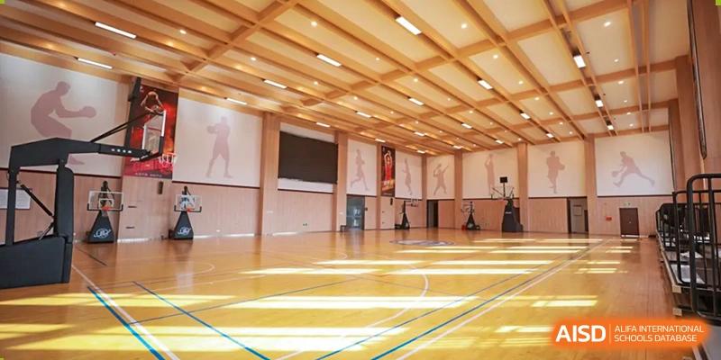 An interior view of a bright, well-maintained indoor gymnasium with a polished wooden floor and high ceiling, featuring several basketball hoops at Hefei Xinhua Academy.