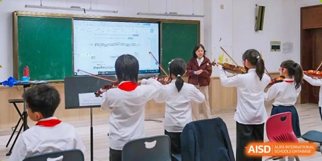 Inside a brightly lit classroom, two young students stand with their backs to the camera, actively practicing the violin while utilizing a large digital smart board displaying sheet music for their lesson at Hefei Xinhua Academy.