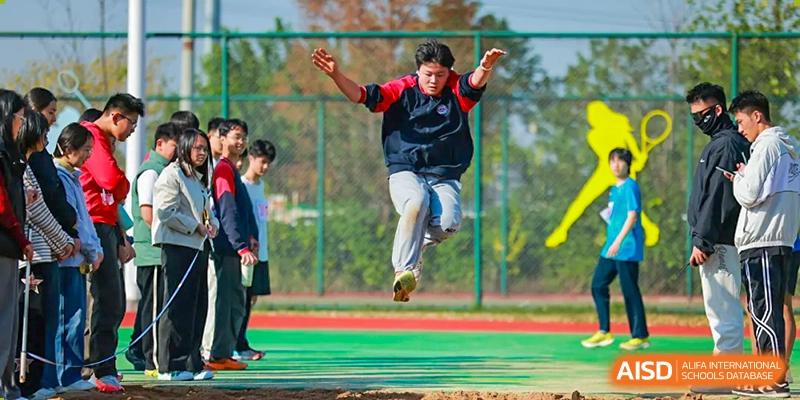 A focused male student is captured mid-air during a competitive long jump attempt on the school's sports field, surrounded by observing classmates cheering him on during an exciting outdoor sports day at Hefei Xinhua Academy.