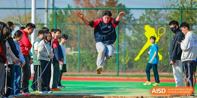 A focused male student is captured mid-air during a competitive long jump attempt on the school's sports field, surrounded by observing classmates cheering him on during an exciting outdoor sports day at Hefei Xinhua Academy.