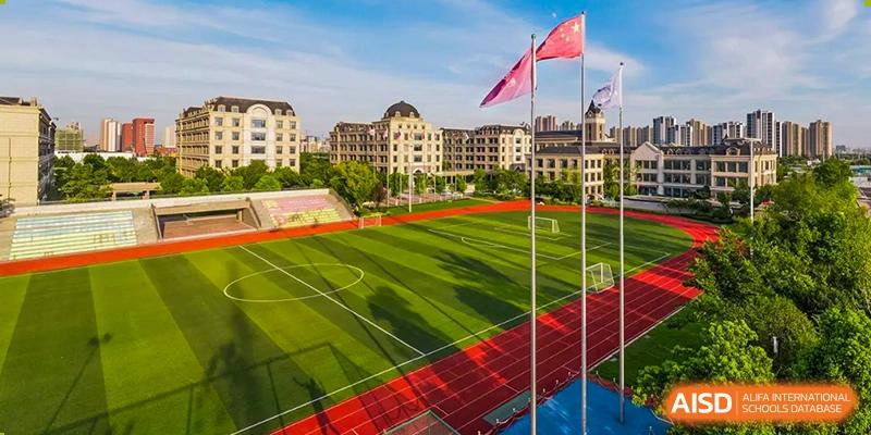 An elevated wide shot showing the well-maintained outdoor red running track and green football field, with school buildings visible in the background at Hefei Xinhua Academy.