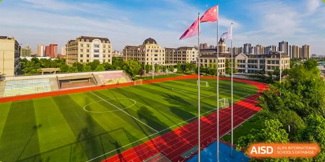 An elevated wide shot showing the well-maintained outdoor red running track and green football field, with school buildings visible in the background at Hefei Xinhua Academy.
