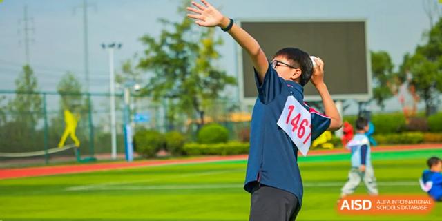 A student wearing a blue sports shirt and a numbered bib is captured throwing an object, possibly a javelin or shot put, during a field event at Hefei Xinhua Academy.