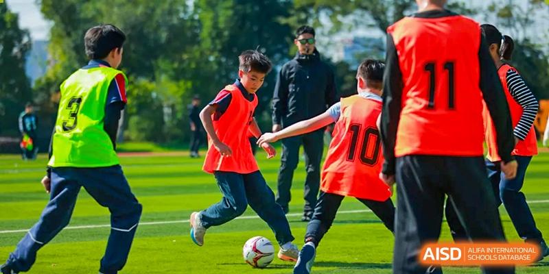 Two students in orange jerseys are actively engaged in a competitive game of soccer on the grass pitch during an outdoor sports lesson at Hefei Xinhua Academy.
