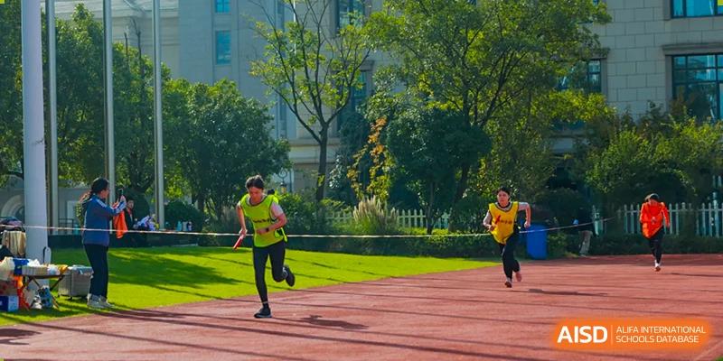 Students are running a race with full effort on the red track during a track and field competition, with trees and spectators in the background at Hefei Xinhua Academy.