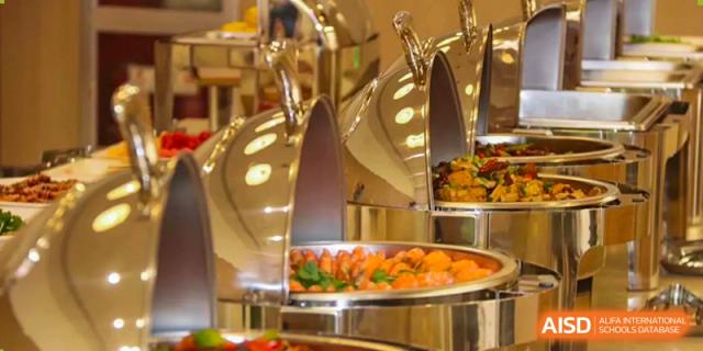 A close-up view of shiny silver chafing dishes filled with prepared buffet food, including colorful vegetables and shrimp, is shown at Hefei Xinhua Academy.