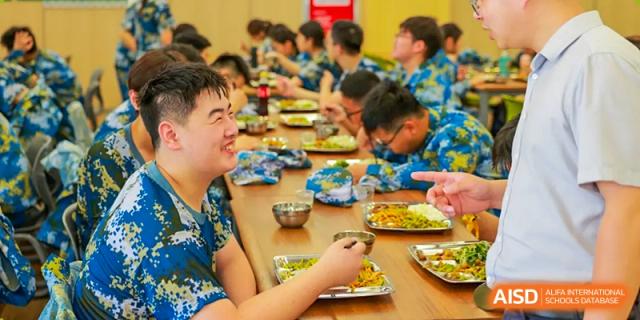 A group of students wearing matching blue and yellow patterned shirts are happily eating their lunch together at a long wooden table at Hefei Xinhua Academy.