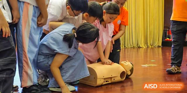 Students are gathered around a training mannequin, practicing CPR compressions and first aid techniques under the supervision of an instructor at Hefei Xinhua Academy.
