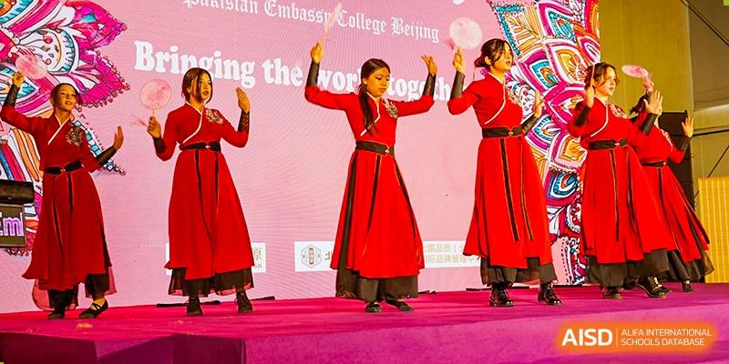 Three students from Pakistan Embassy College Beijing performing a traditional dance on stage in flowing red dresses.