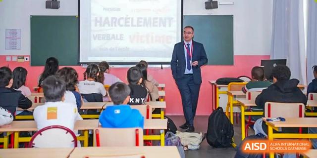 Man in a suit, identified as the Proviseur, du Lycée Français International de Pékin, presenting a lesson on "HARCÈLEMENT VERBALE VICTIME" to a class of young students seated at desks in a brightly lit classroom.