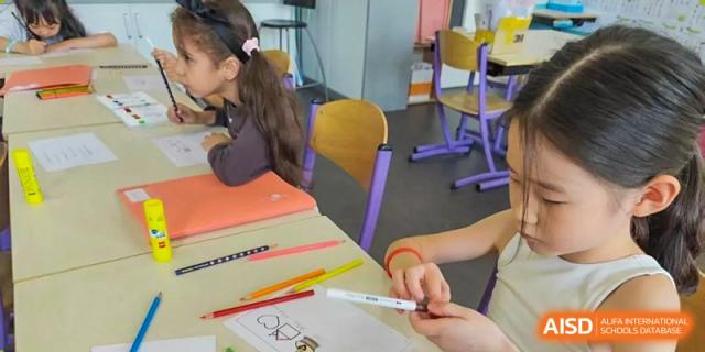 Close-up on a student's hands drawing a picture with a marker at a desk, surrounded by colored pencils in a classroom setting at the Proviseur, du Lycée Français International de Pékin, focusing on hands-on learning.