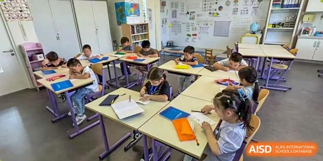 Several young students at the Proviseur, du Lycée Français International de Pékin, intently writing in notebooks while seated at double desks in a classroom with educational posters displayed on the wall behind them.